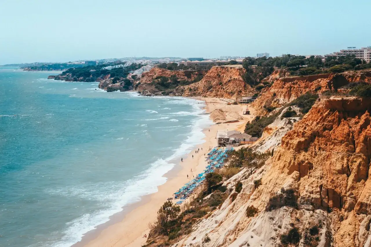 TUI Blue Falesia: Türkisfarbenes Meer trifft auf ockerfarbene Felsen. Ein Strand mit Liegen und ein paar Häusern lädt zum Entspannen ein.