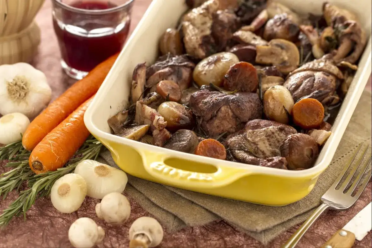 Un plat de coq au vin mijot&eacute;, avec des morceaux de poulet, des lardons, des champignons, des oignons et des carottes, servi dans un plat jaune.