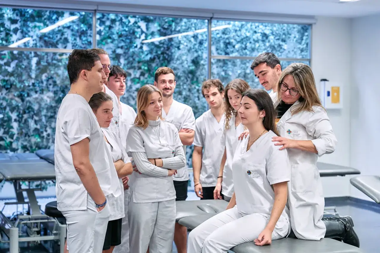 Estudiantes de fisioterapia aprenden t&eacute;cnicas de palpaci&oacute;n en un aula. Un grupo diverso de j&oacute;venes, con batas blancas, observa atentamente.