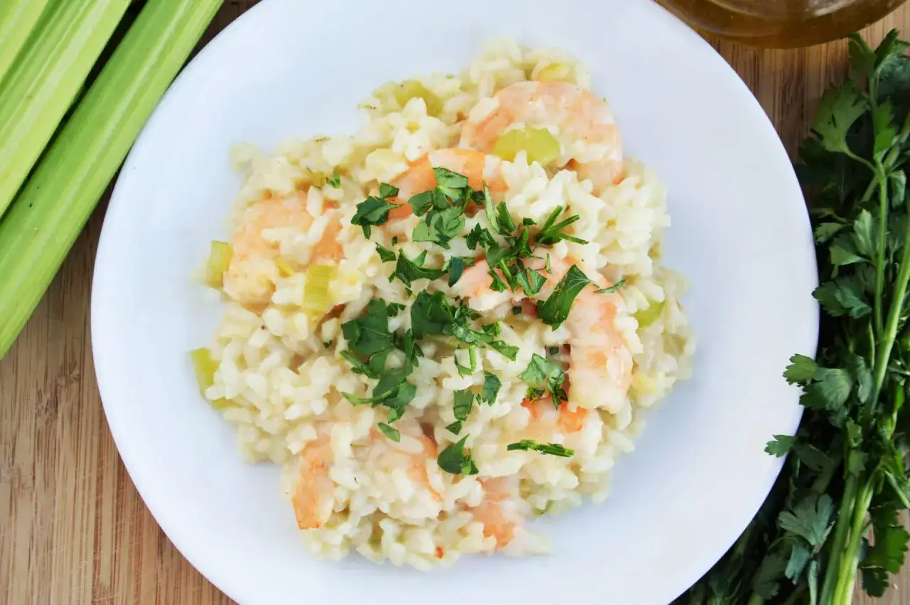 Kremowe risotto z krewetkami, ozdobione natką pietruszki, obok łodyg selera i oliwy.