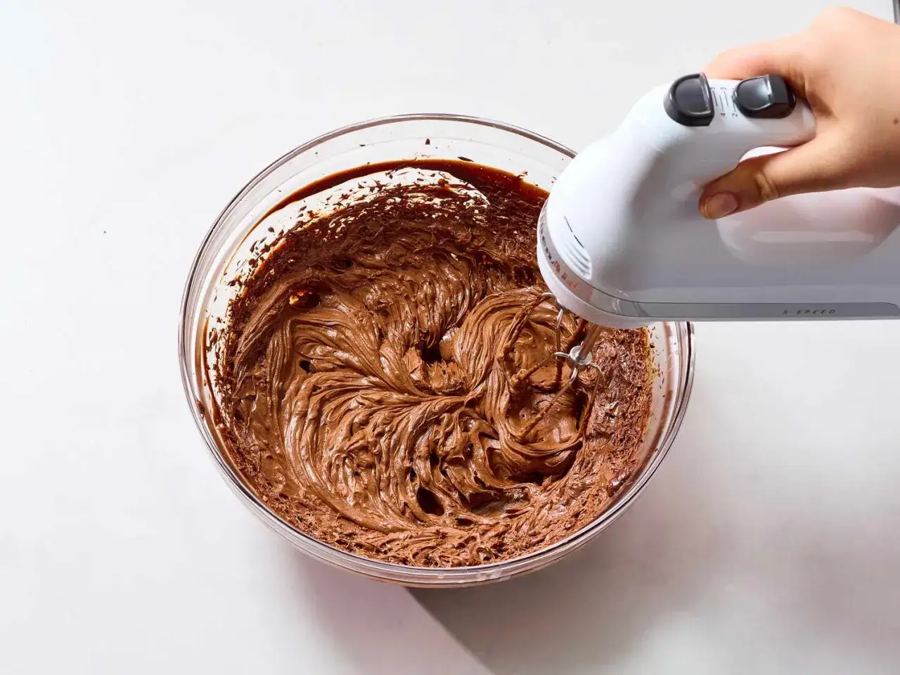 Eine Hand mixt mit einem Handmixer eine cremige Schoko-Ganache in einer Glassch&uuml;ssel.