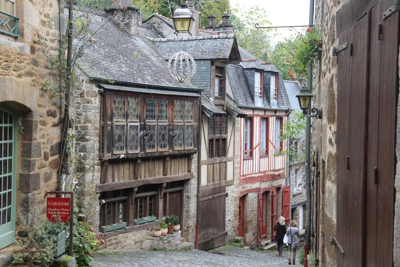 Cobblestone street in Dinan, France, lined with historic stone and timber-framed buildings. Two women walk away from the camera.