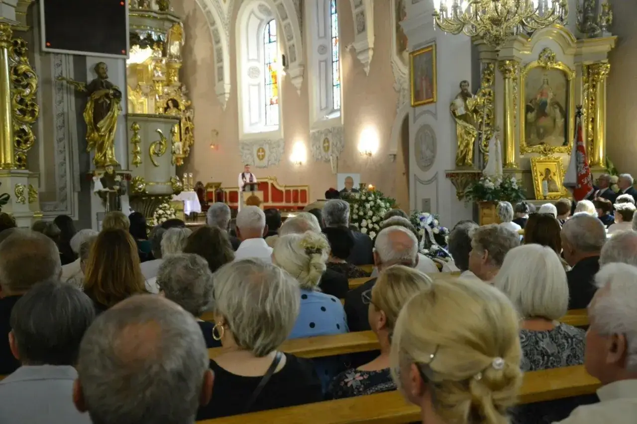 Ksiądz odczytuje pożegnanie zmarłego. W kościele zgromadzeni wierni, przy ołtarzu kwiaty i portret zmarłego.