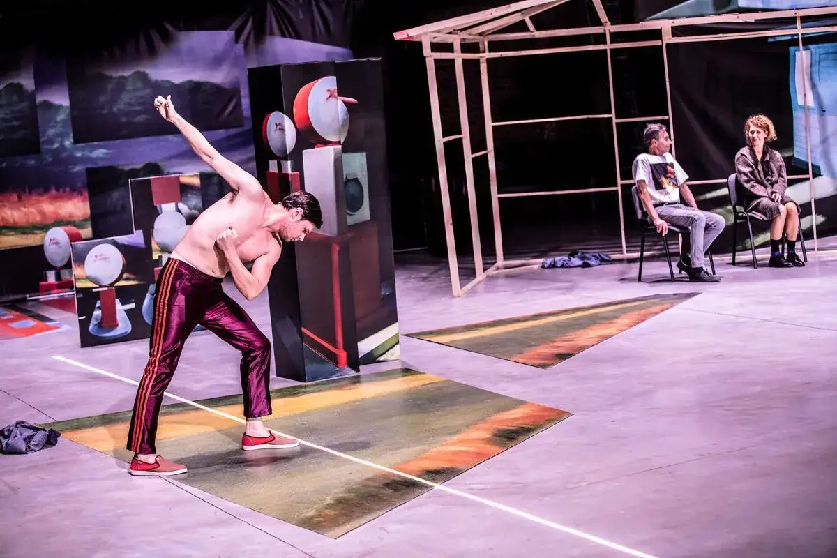 Aktorzy Teatru Polskiego we Wrocławiu na scenie. Mężczyzna w błyszczących spodniach w dynamicznej pozie, obok scenografia.
