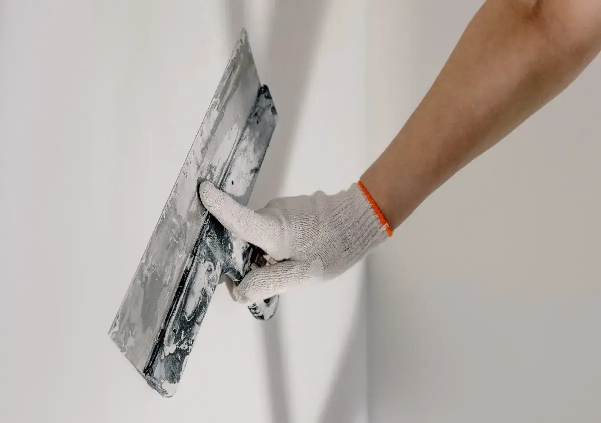 Mano enguantada aplica mortero sobre pared blanca con llana. Se puede aplicar mortero sobre pintura para renovar superficies.