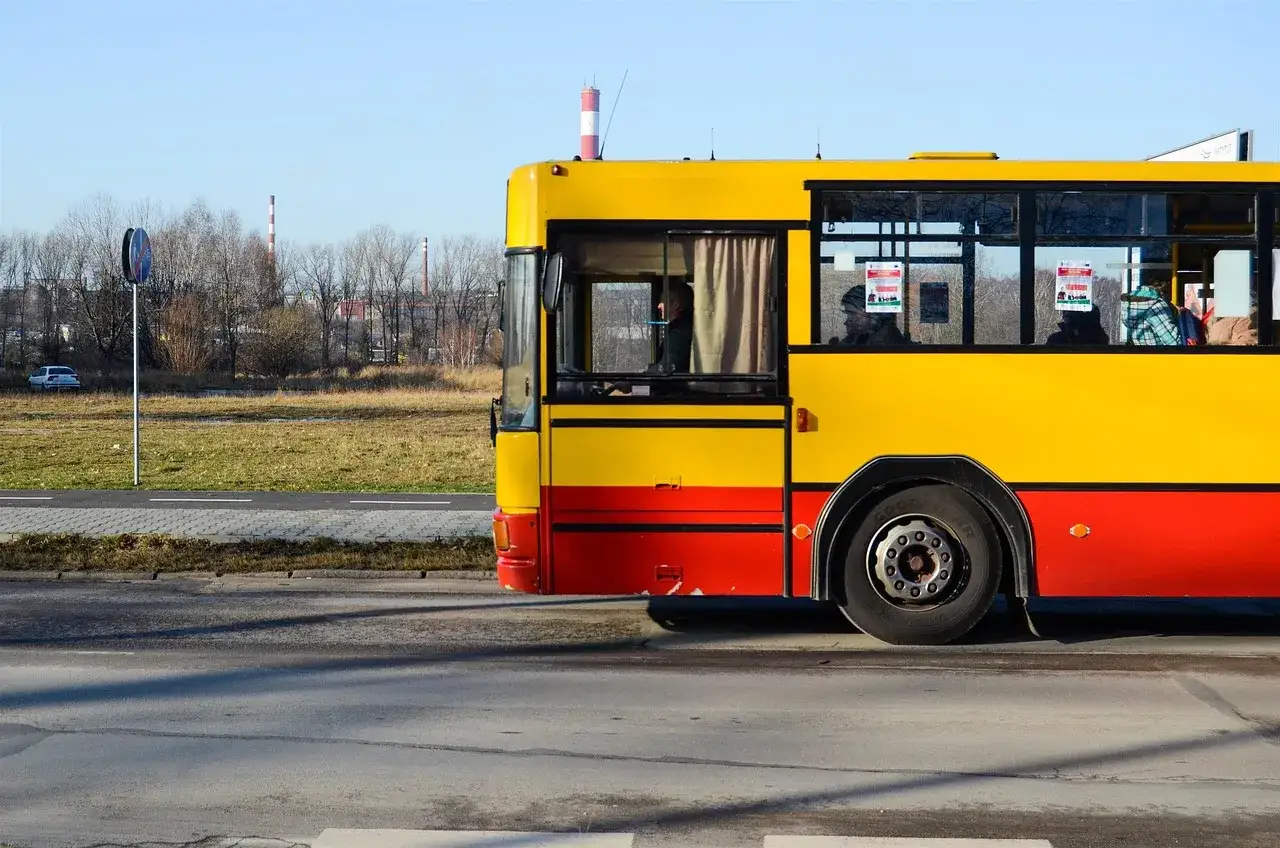 Żółto-czerwony autobus jedzie drogą. W tle widać drzewa i kominy, jakby wyjeżdżał z **puszczy białowieskiej**.