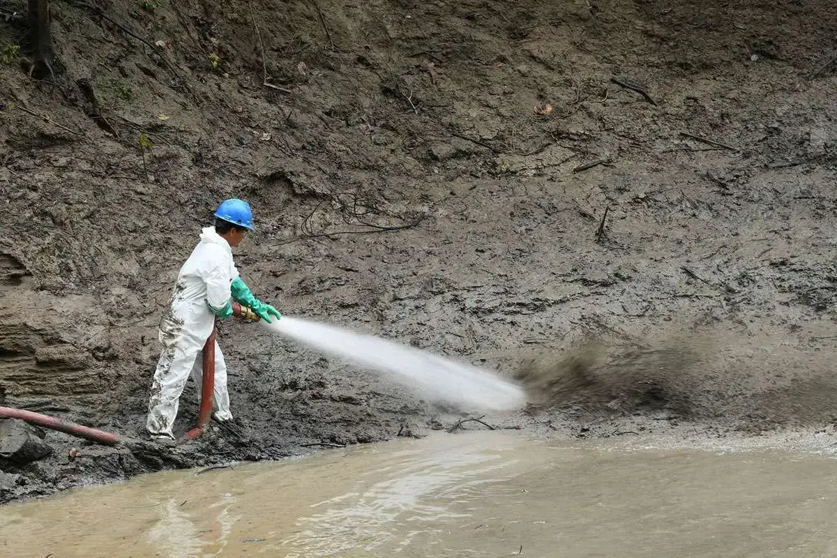 Trabajador con traje protector realiza remediación de suelos, lavando la tierra con una manguera.