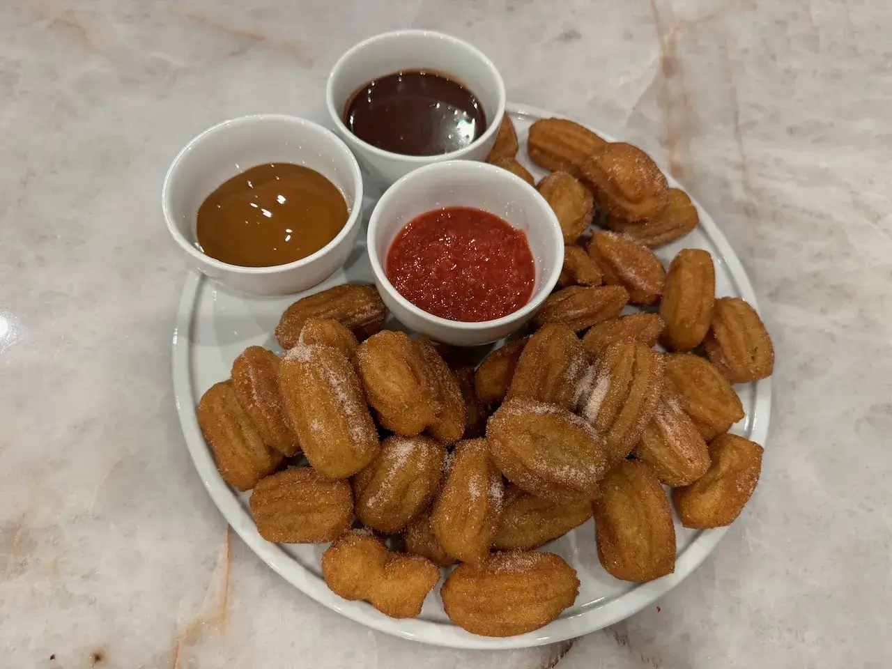 En tallrik med sockerstr&ouml;sslade churros, serverade med tre dipps&aring;ser: choklad, kolas&aring;s och jordgubbssylt. Perfekt f&ouml;r att njuta av vad &auml;r churros!