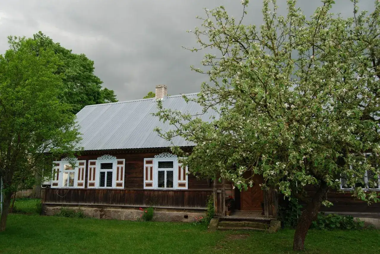 Drewniany dom z ozdobnymi okiennicami, kwitnąca jabłoń i zieleń wokół. Urokliwe studzianki w tle.