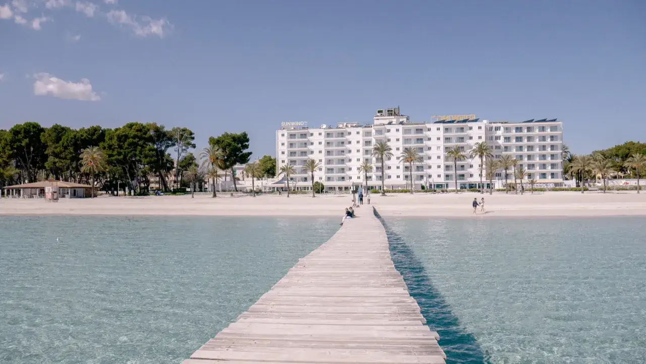 Drewniany pomost prowadzi do piaszczystej plaży Playa de Muro z białym hotelem i palmami w tle.