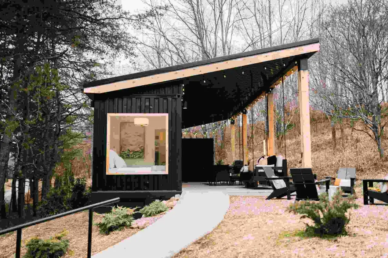 Una moderna casa en un container, con un porche cubierto y asientos al aire libre, rodeada de árboles.