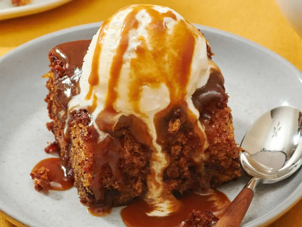 En bit saftig sticky toffee pudding med vaniljglass och kolasås som rinner ner. En sked ligger bredvid.