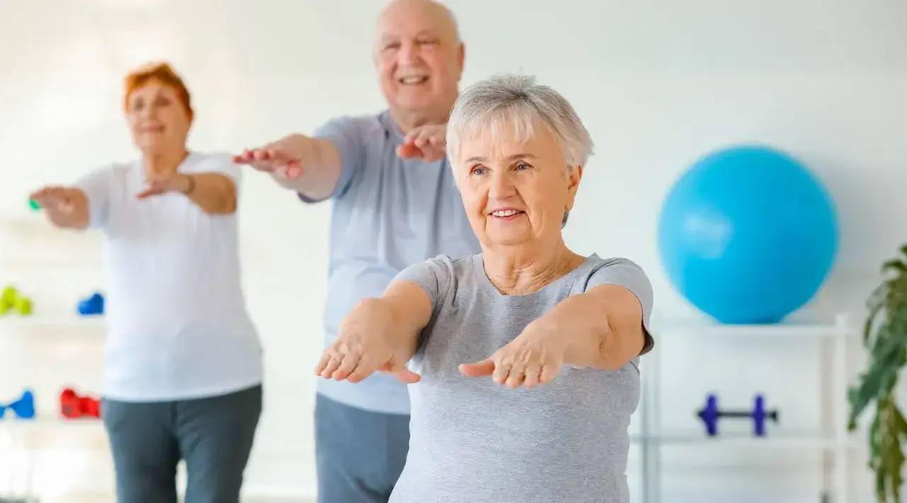 Trois seniors souriants pratiquent la gym douce, bras tendus, dans une salle lumineuse.