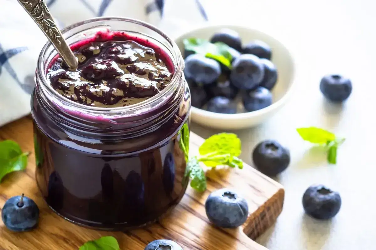 Vasetto di marmellata di mirtilli fatto in casa, con mirtilli freschi e foglie di menta su un tagliere di legno.