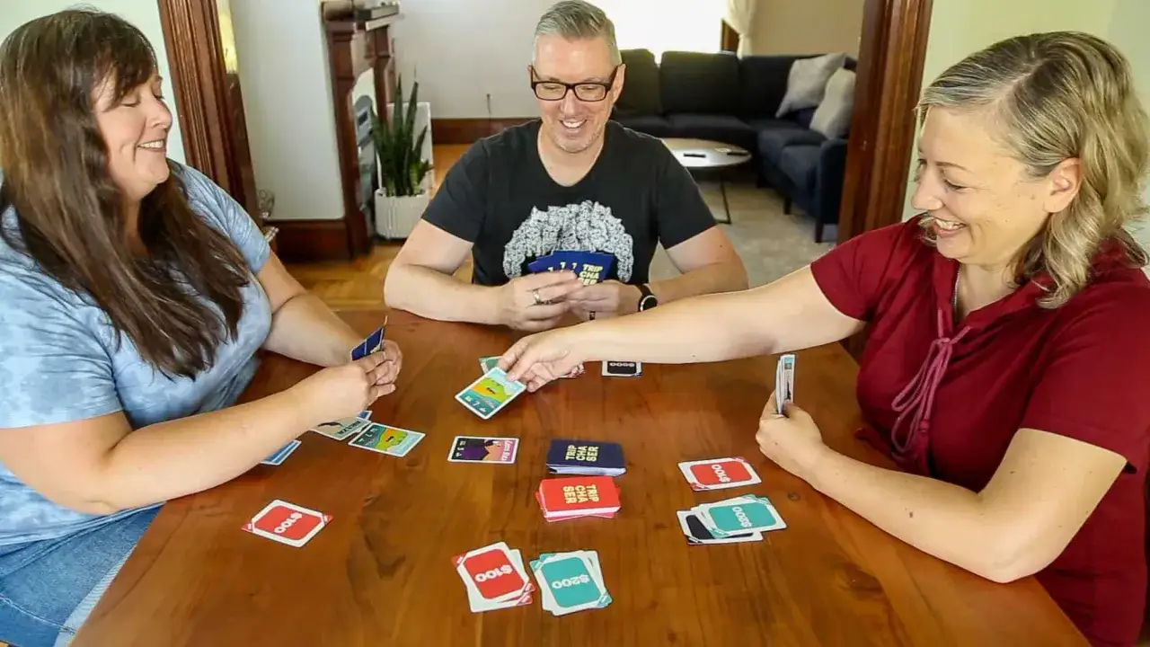 Drei lachende Erwachsene spielen Karten am Holztisch. Bunte Karten liegen aus, perfekt für Reisespiele für Erwachsene.