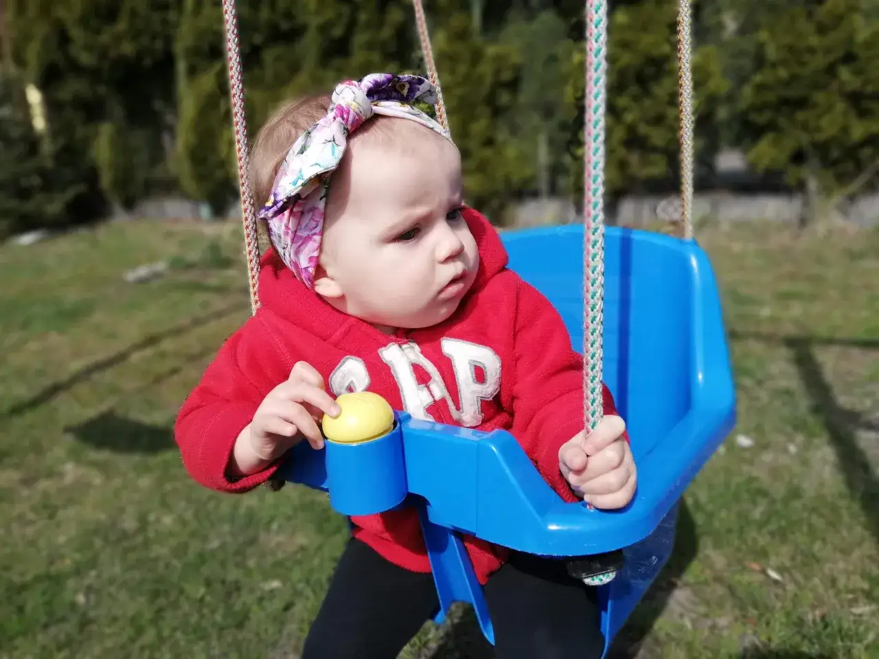 Uśmiechnięte 15 miesięczne dziecko w czerwonej bluzie z kapturem i kolorowej opasce na głowie bawi się na niebieskiej huśtawce.