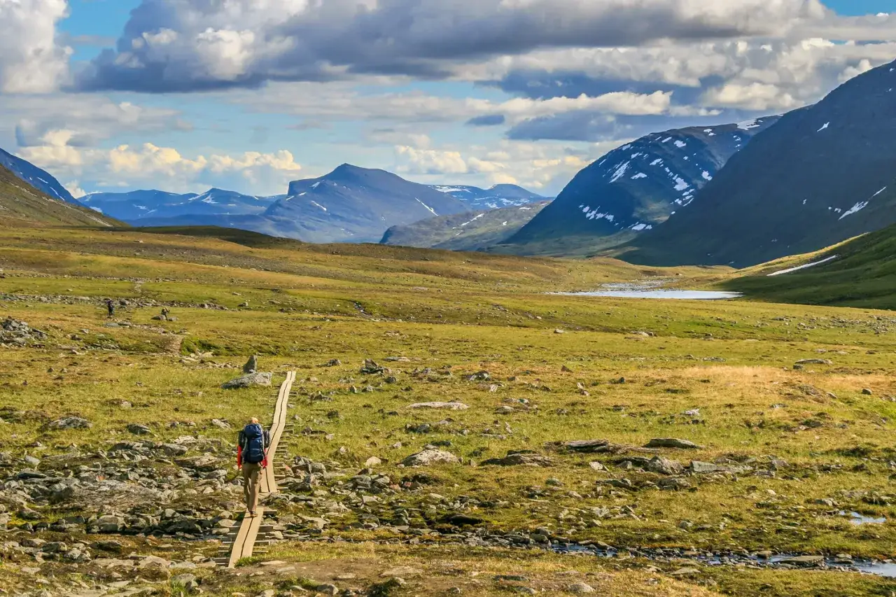 Vandra kungsleden i ett storslaget fjällandskap med en ensam vandrare på en spång över en grön dal.