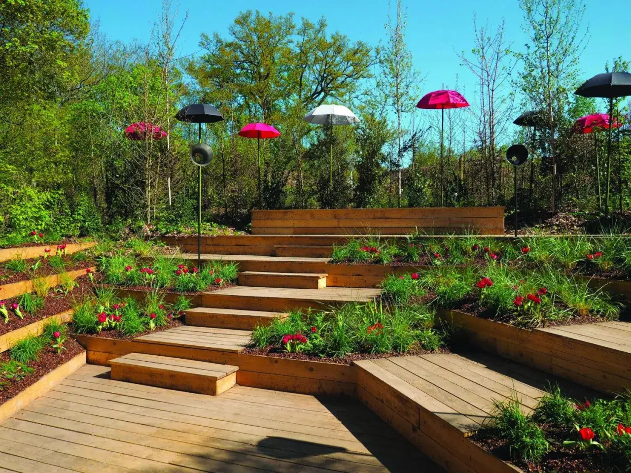 Un jardin am&eacute;nag&eacute; avec des terrasses en bois et des parterres de fleurs. La profondeur du marche escalier ext&eacute;rieur est accentu&eacute;e par des parasols color&eacute;s.