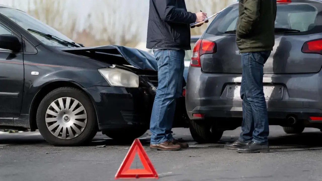 Dwaj mężczyźni spisują szkody po wypadku. Uszkodzone samochody i trójkąt ostrzegawczy sugerują, że może to być początek sprawy o karę za spowodowanie wypadku z uszczerbkiem na zdrowiu.