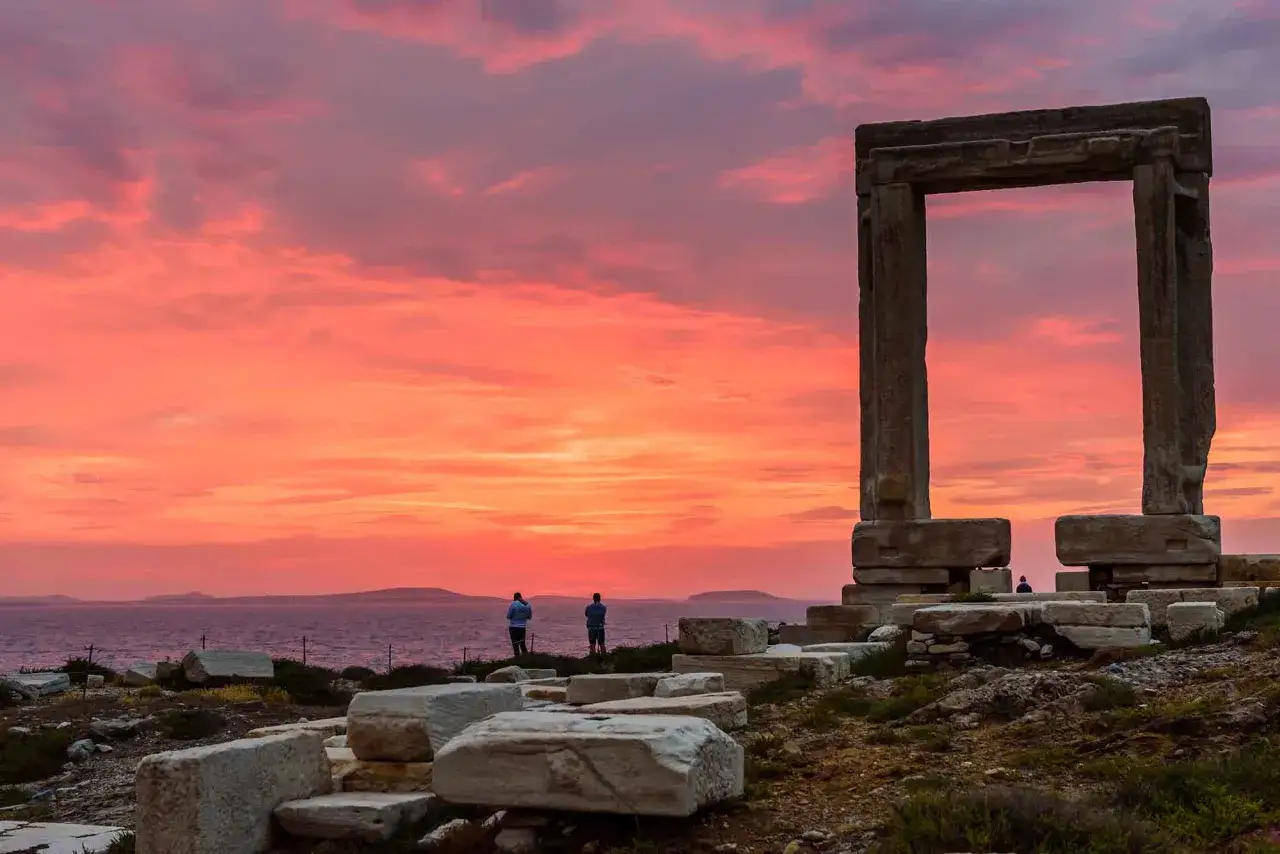 Ancient ruins on Naxos, Greece, stand silhouetted against a vibrant sunset sky. Two figures gaze out at the sea.