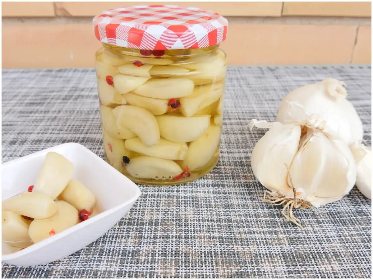 Aglio sott'olio fatto in casa da Benedetta, con spicchi dorati e peperoncino in un barattolo e una ciotolina, accanto a bulbi freschi.