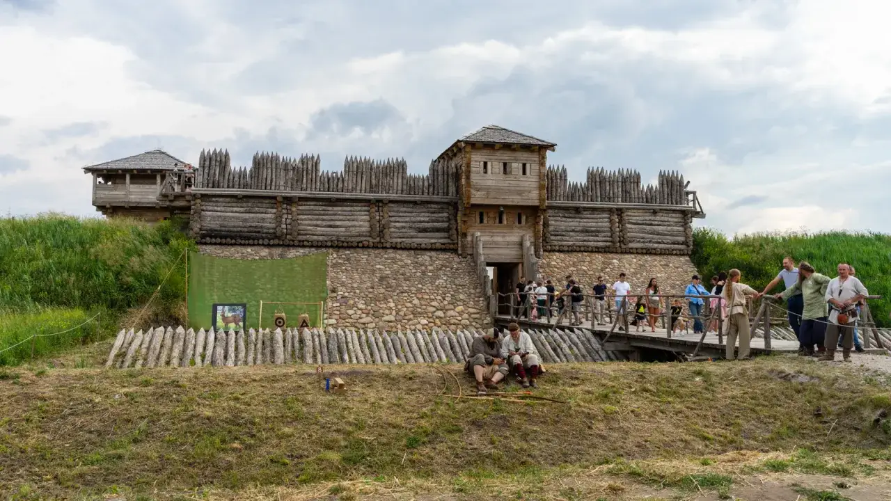 Rekonstrukcja grodziska w Tumie z drewnianymi wieżami i kamiennym murem. Ludzie spacerują po moście, a dwaj mężczyźni w strojach historycznych siedzą na trawie.