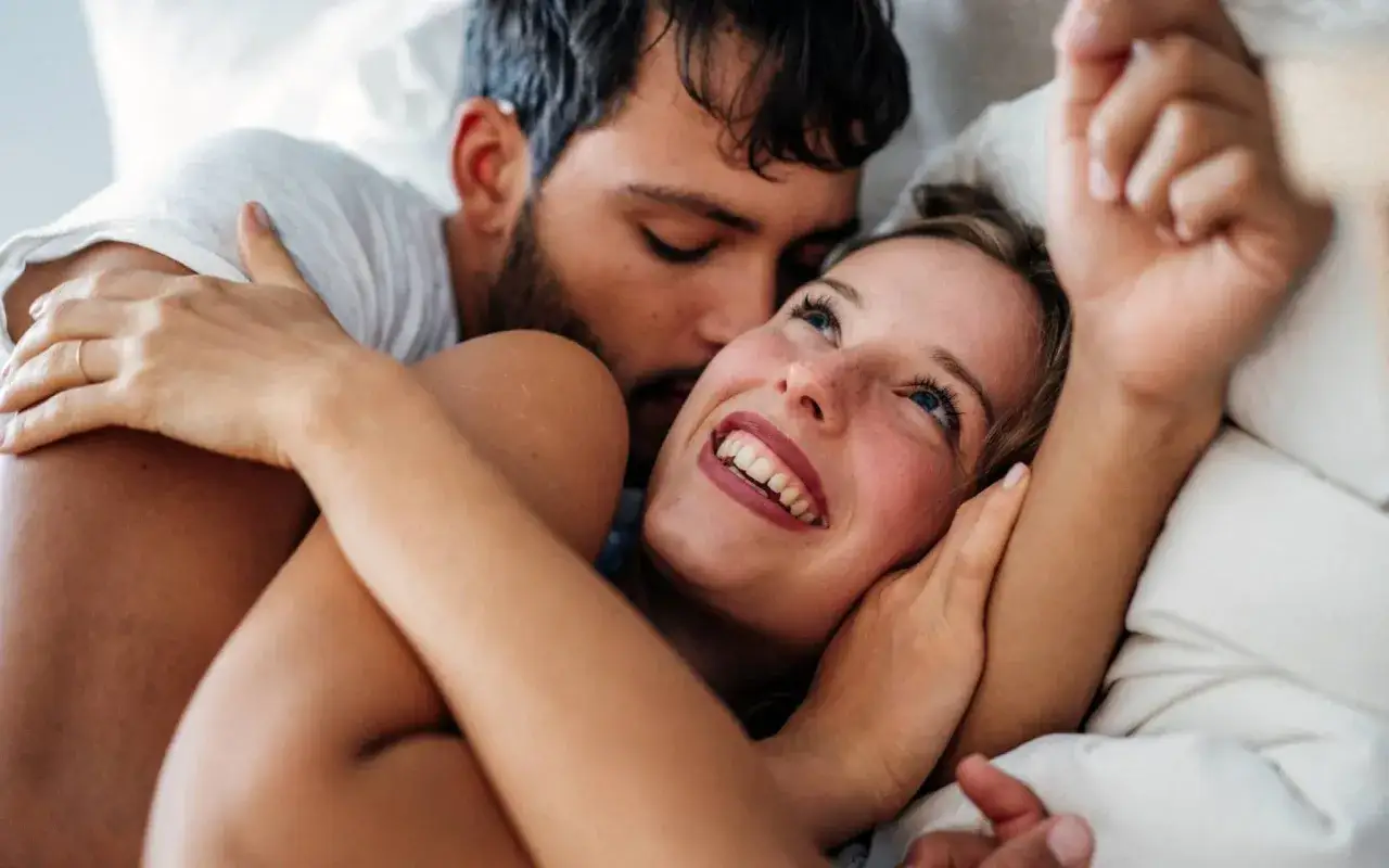 Ein Mann umarmt eine Frau im Bett. Sie lächelt ihn an. Manchmal hat man keine Lust auf Sex in der Schwangerschaft, aber Nähe ist trotzdem schön.
