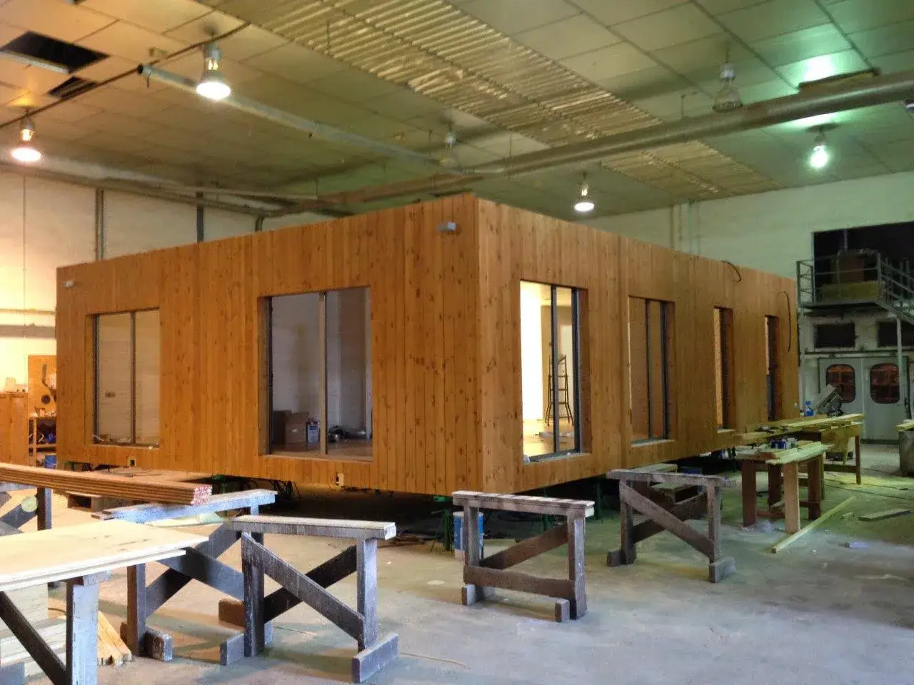 Une maison modulable en bois, avec de grandes fenêtres, est en cours de construction dans un atelier.