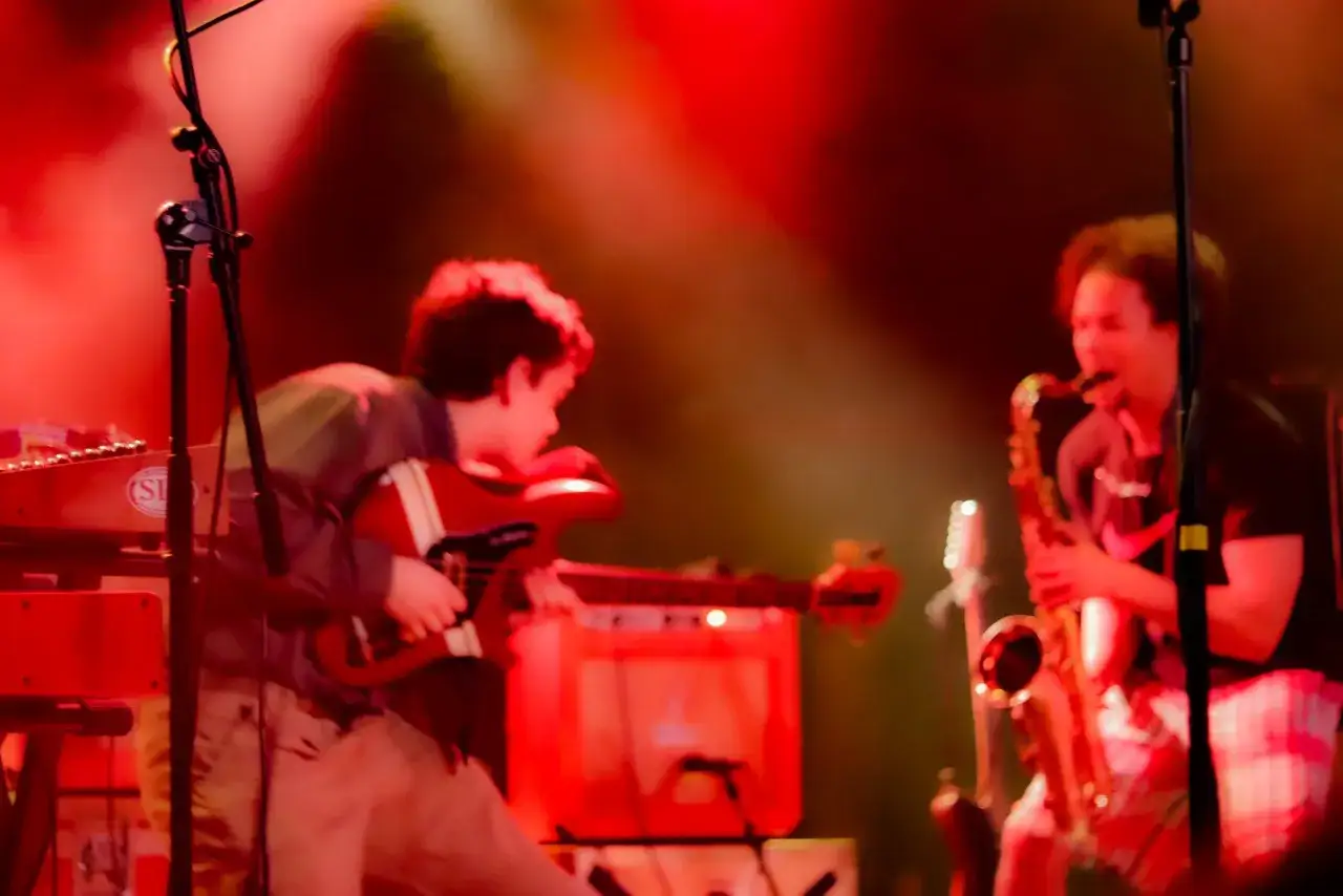 Un groupe de musique, black midi, joue sur scène sous des lumières rouges. Un guitariste et un saxophoniste sont visibles.