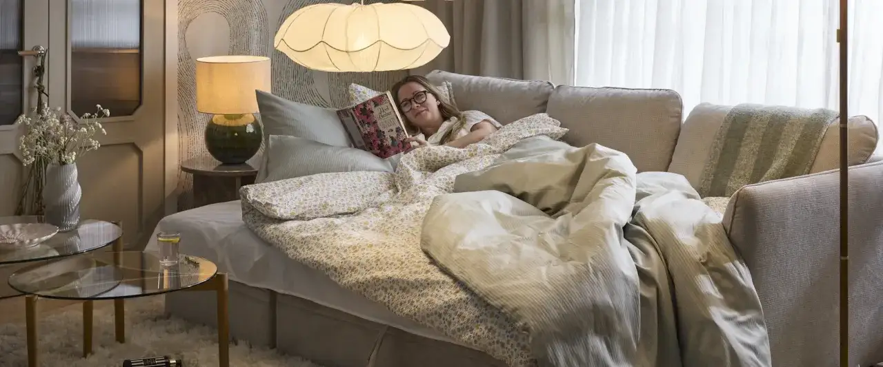 En kvinna läser en bok på en bekväm, ergonomisk soffa, omgiven av mjuka täcken och kuddar.