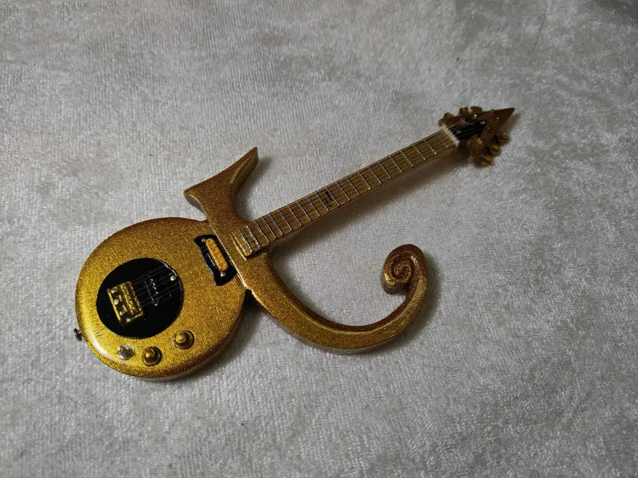 Złota gitara w kształcie symbolu Prince'a, błyszcząca i ozdobna, leży na białym, puszystym tle.