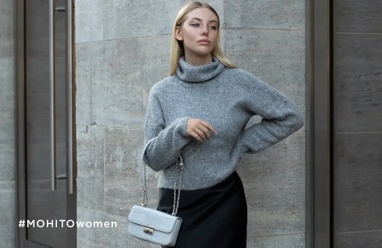 Kobieta w szarym golfie i czarnej spódnicy, z szarą torebką na łańcuszku. Stylizacja w duchu moda quiet luxury.