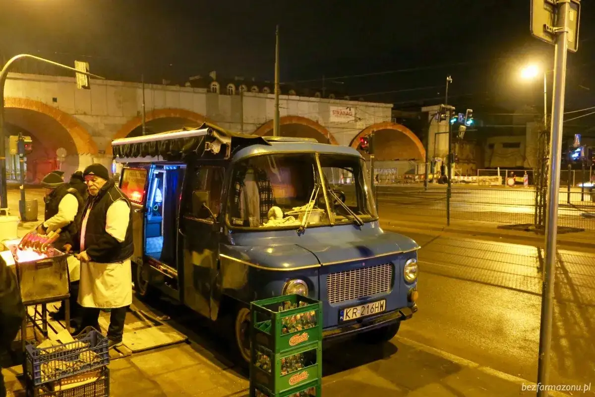 Nocny sprzedawca przygotowuje pyszne kiełbaski z nyski. W tle widać łuki wiaduktu i miejskie światła.