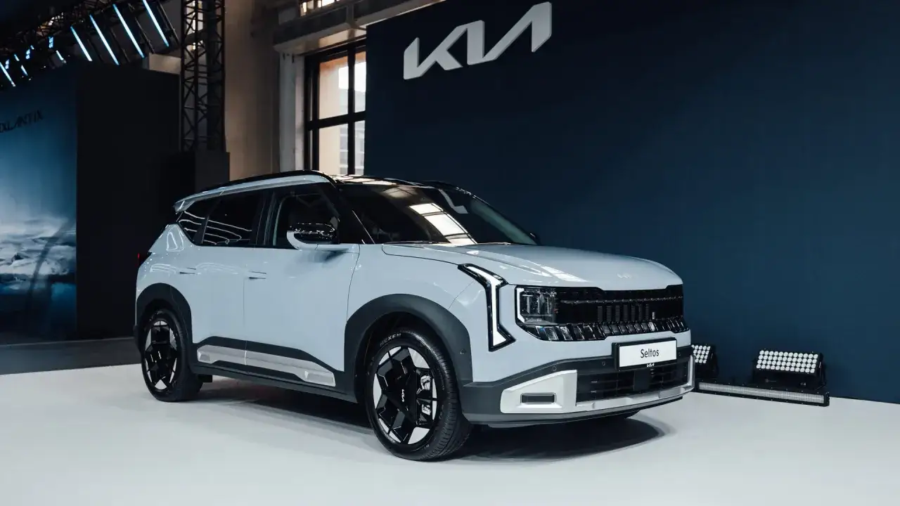 Nowy, jasnoszary suv Kia Seltos prezentuje się na tle ciemnoniebieskiej ściany z logo marki.
