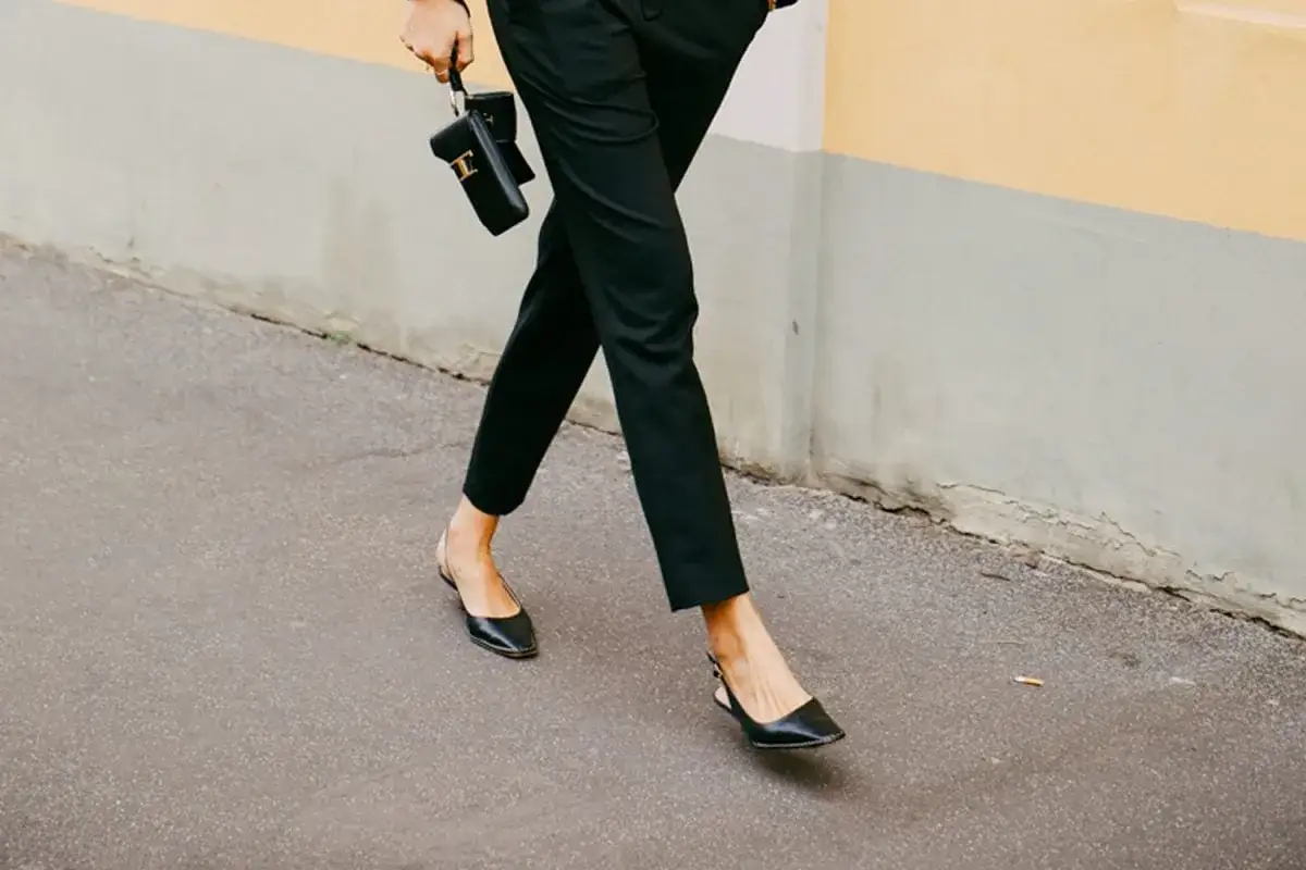 Une femme élégante marche dans la rue, portant un pantalon noir et des chaussures ballerines noires. Elle tient un petit sac noir.