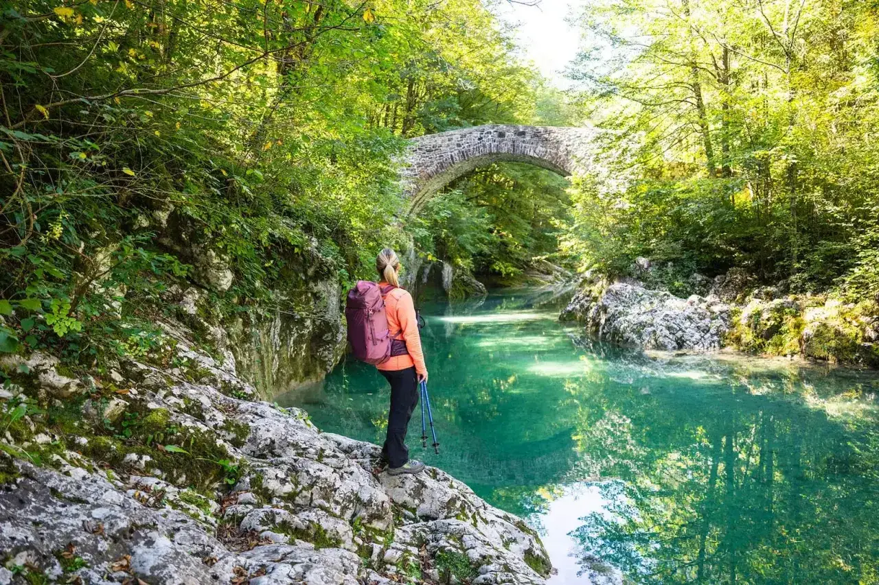 Wanderin am Juliana Trail entlang eines smaragdgr&uuml;nen Flusses, mit einer Steinbr&uuml;cke im Hintergrund.