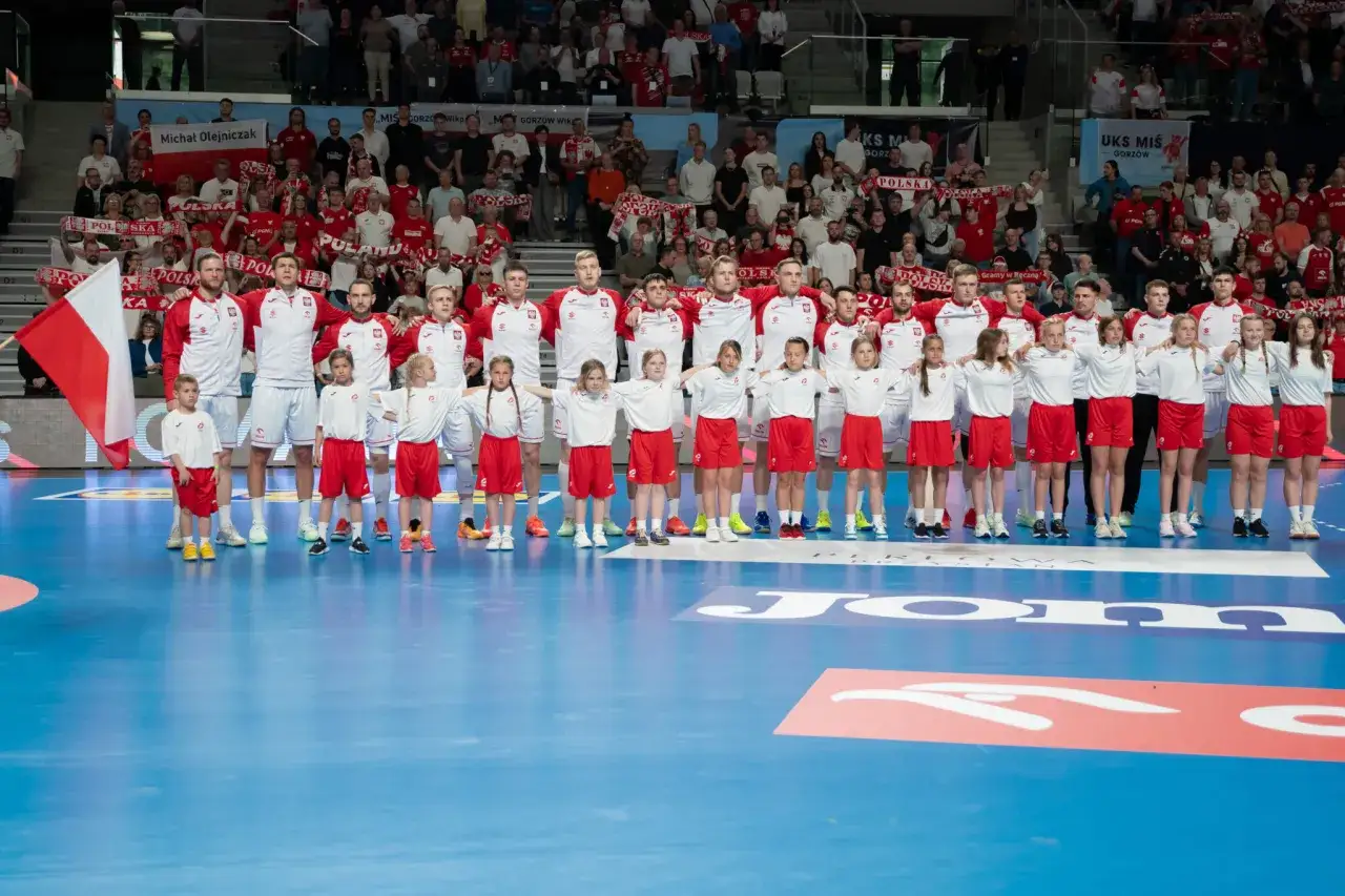 Drużyna piłka ręczna w strojach biało-czerwonych, z dziećmi, na tle publiczności.