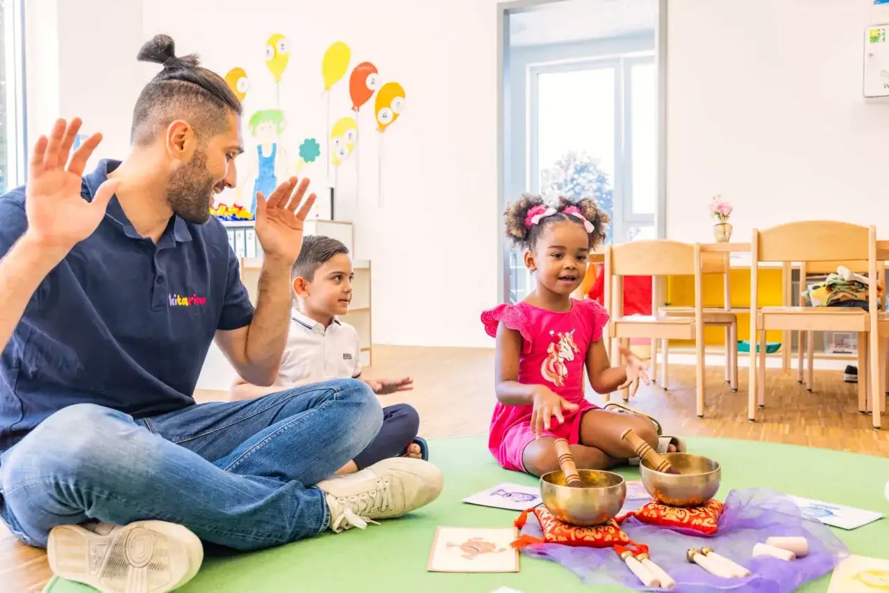 Ein Mann mit Bart und Dutt, ein Junge und ein M&auml;dchen sitzen auf einer gr&uuml;nen Matte. Sie machen eine Klangschalen-&Uuml;bung, ein situationsorientierter Ansatz in der Kita.