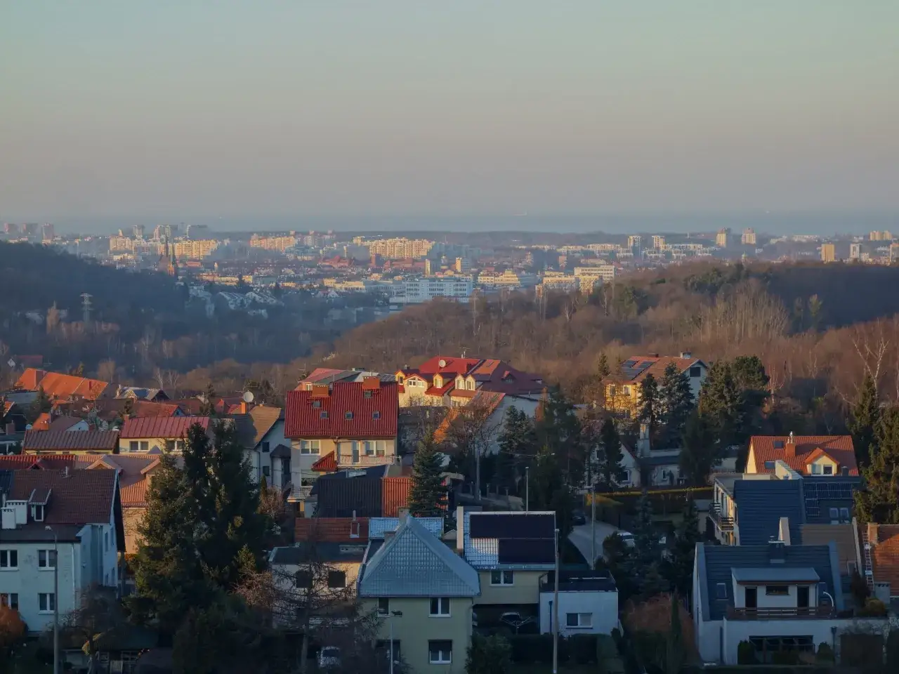 Widok na zabudowania mieszkalne w Gdańsku Suchanino o zachodzie słońca. W oddali widać panoramę miasta i morze.