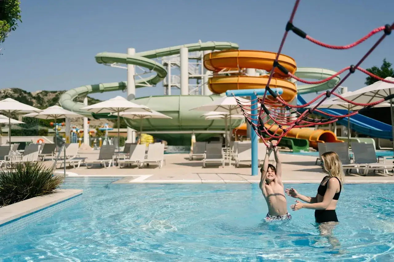 Ein Junge hangelt sich im Pool des Amada Colossos Resort Rhodos, während seine Mutter zusieht. Im Hintergrund bunte Wasserrutschen.