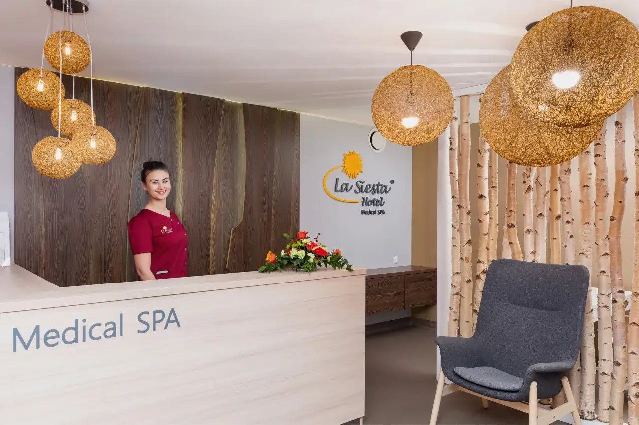 Recepcja La Siesta Hotel Medical SPA. Uśmiechnięta pracownica za ladą, ozdobioną napisem 