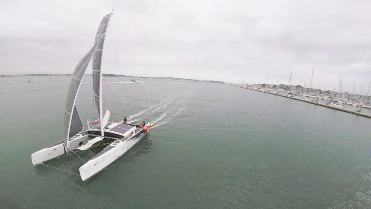 Un catamaran TS3 fend les vagues, ses voiles grises gonflées par le vent. Au loin, un port rempli de voiliers.