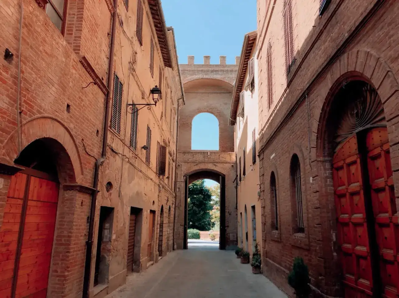 Un vicolo antico con edifici in mattoni rossi e un arco imponente che conduce a un giardino. Sembra un buon convento.