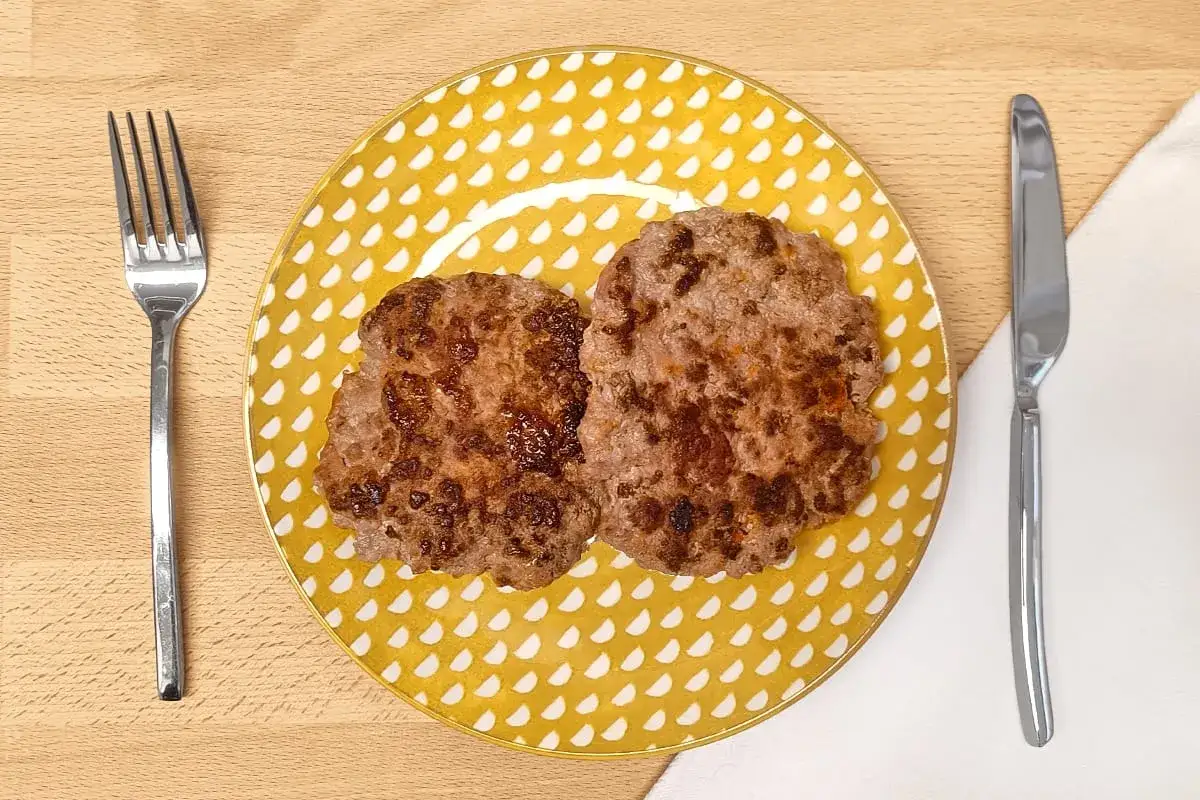 Dos hamburguesas para bebes, doradas y jugosas, servidas en un plato amarillo con puntos blancos. Cubiertos a los lados.