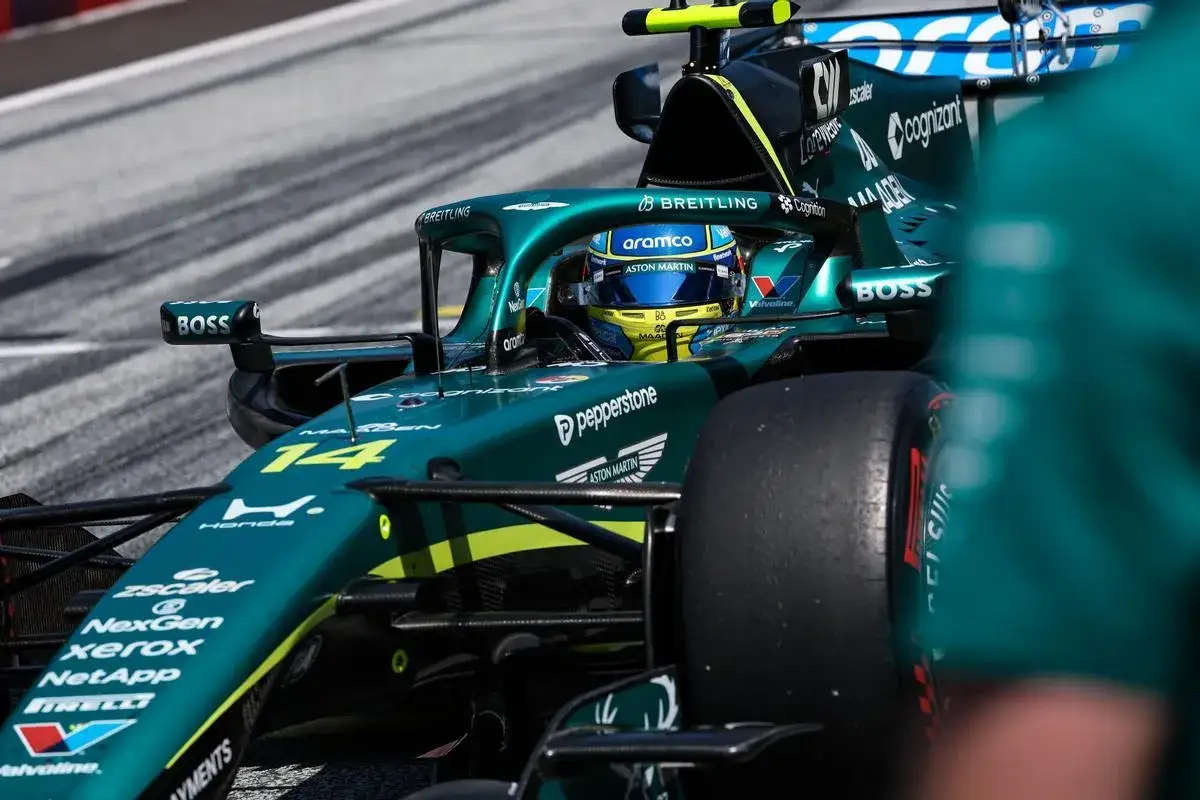 Piloto de Aston Martin F1 con casco azul y amarillo, listo para competir. El coche verde esmeralda luce el número 14.