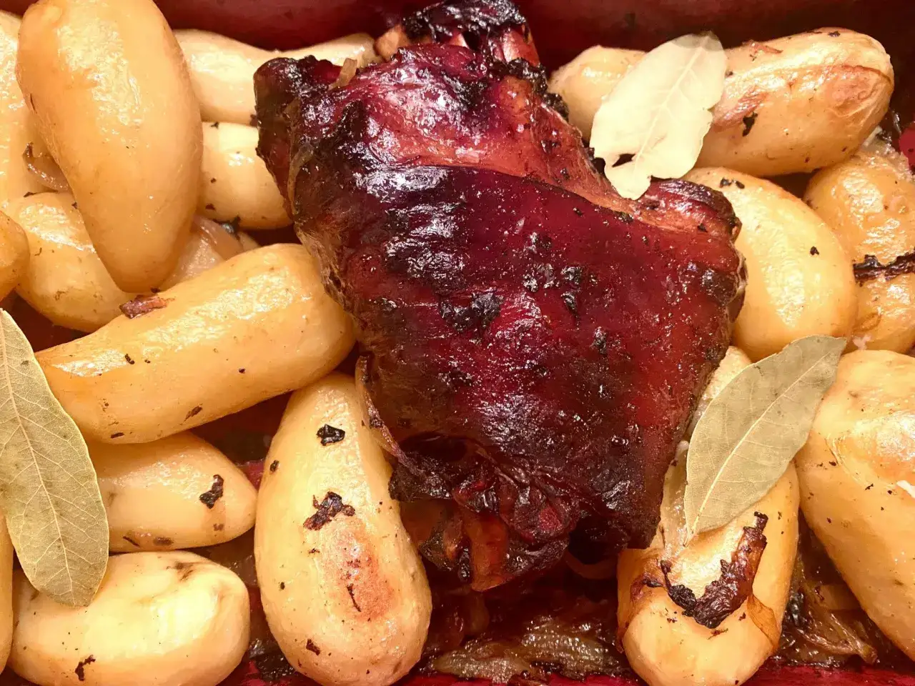 Gros plan sur des kakos de porc rôtis, brillants et caramélisés, entourés de pommes de terre dorées et de feuilles de laurier.