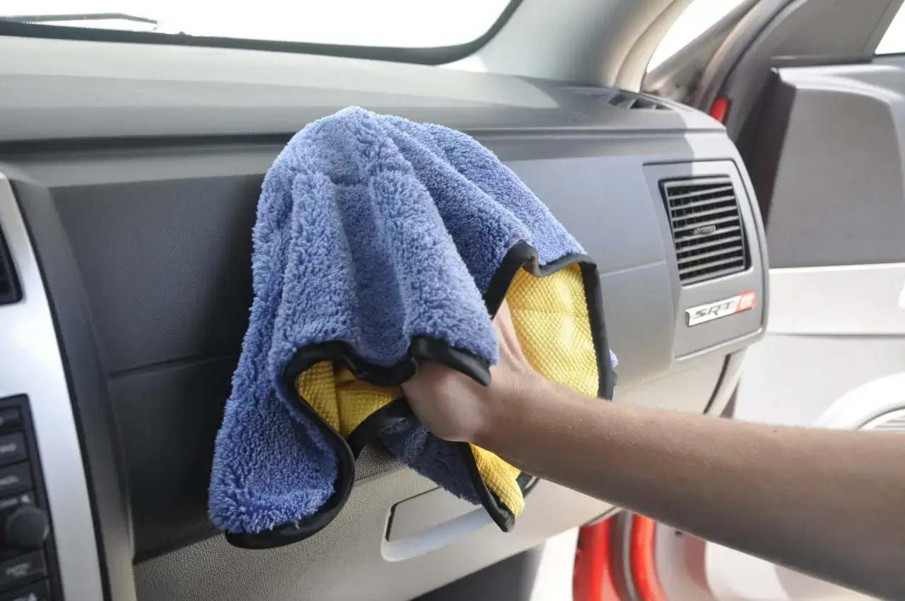 A hand wipes the car's dashboard with a microfiber cloth. Does wax make car shine? This cloth helps maintain that shine.