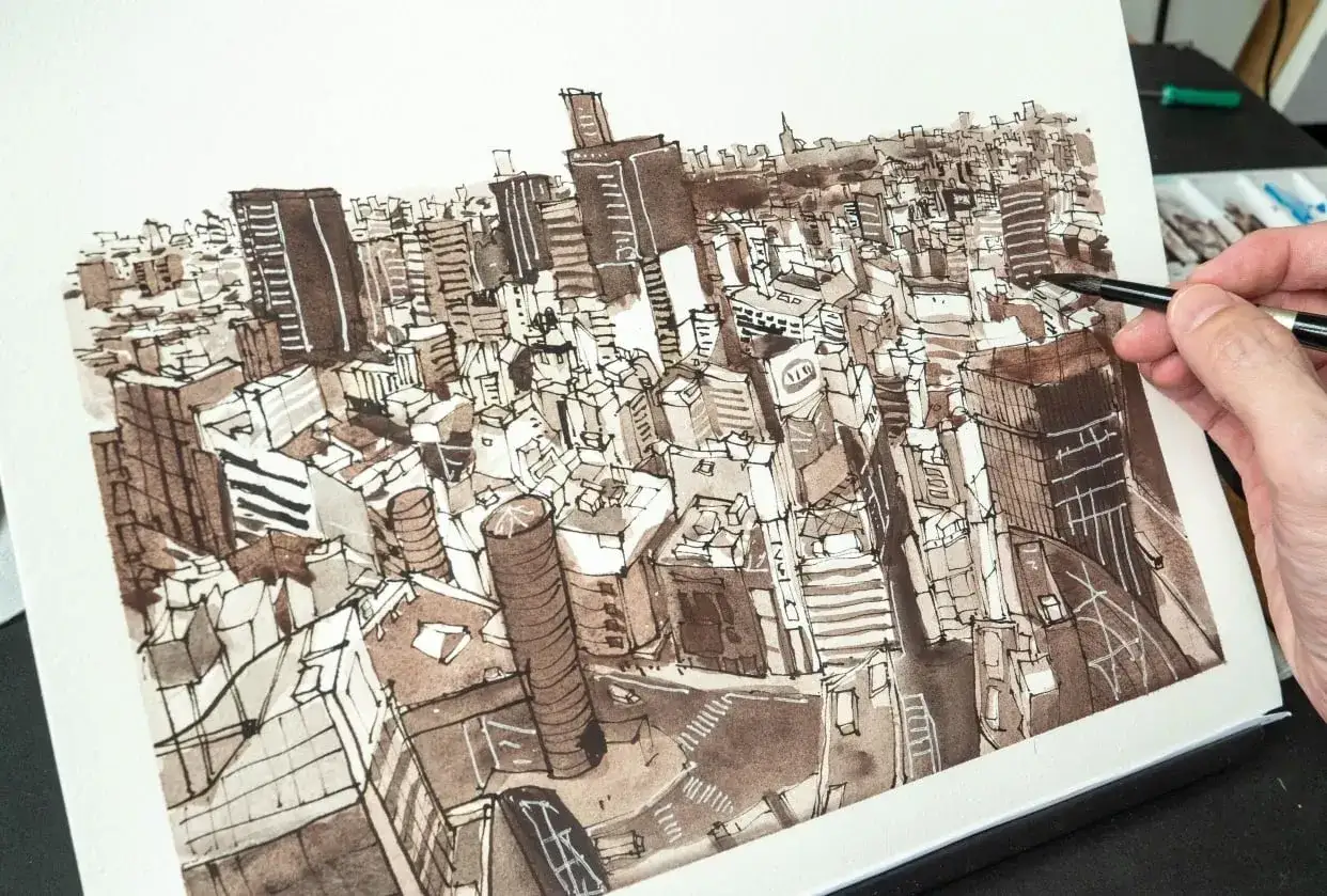 Vue a&eacute;rienne d'une ville dessin&eacute;e &agrave; l'encre, avec des gratte-ciel denses. Au premier plan, une main tient un pinceau, ajoutant des d&eacute;tails &agrave; l'&oelig;uvre.