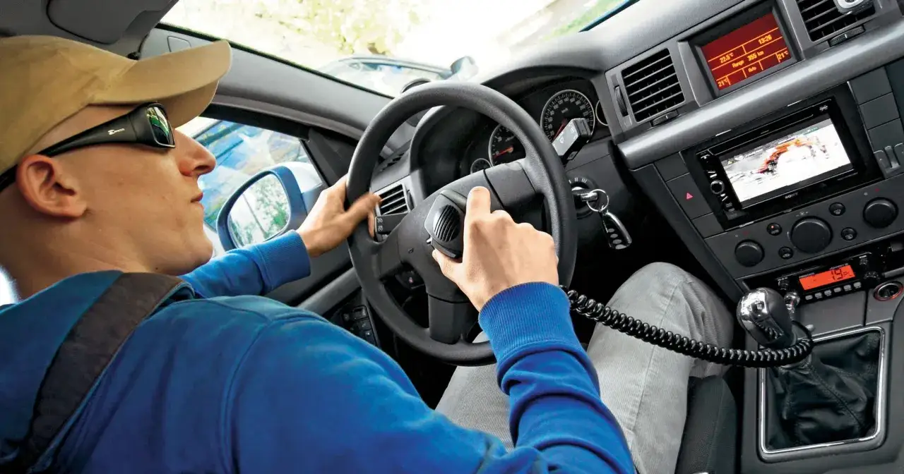 Mężczyzna w czapce i okularach prowadzi auto, trzymając w ręku mikrofon do rejestracji cb. Na desce rozdzielczej widoczny ekran radia.