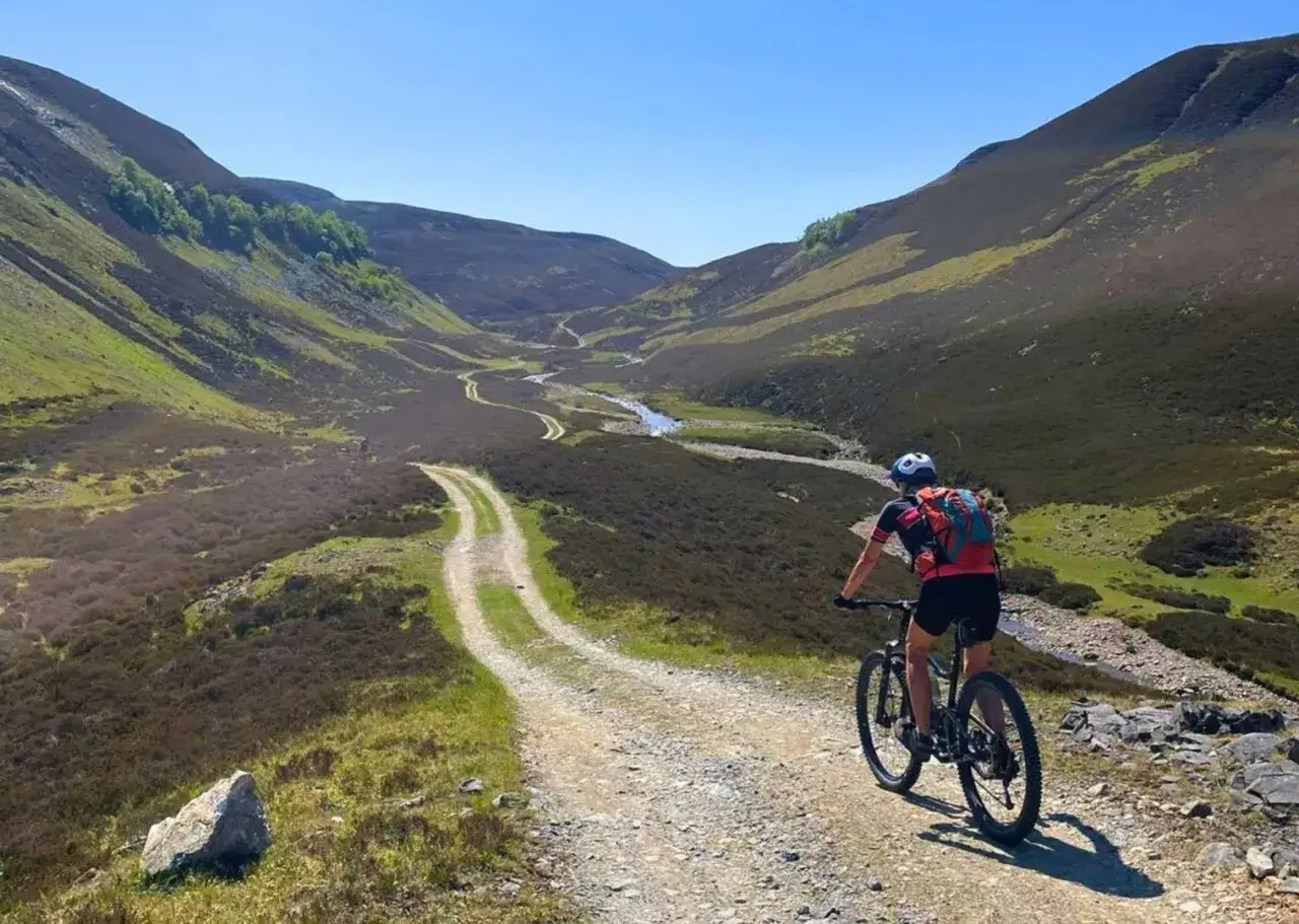 A person enjoys cycling in Scotland, riding a mountain bike on a winding dirt path through a lush, green valley with a stream.